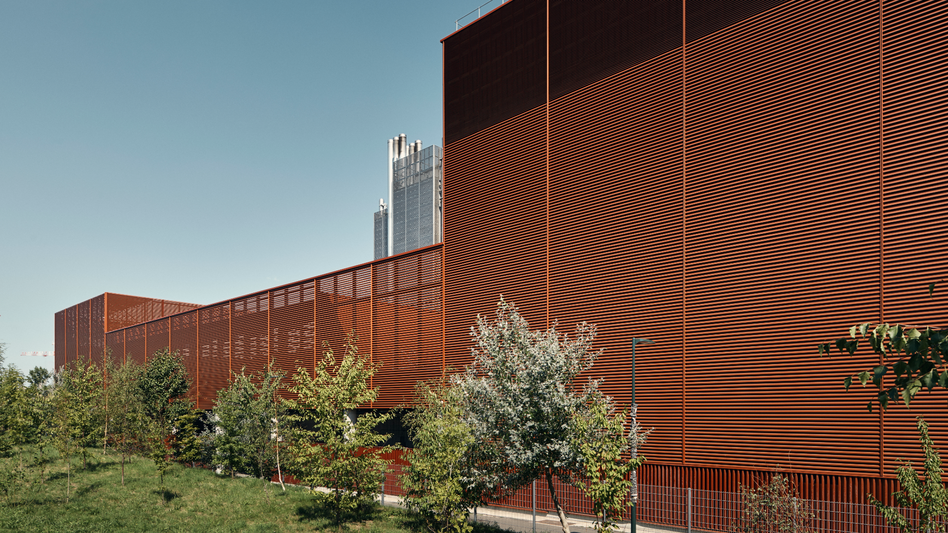 Red corrugated building with horizontal orange-red panels, green trees in front and a clear blue sky behind.