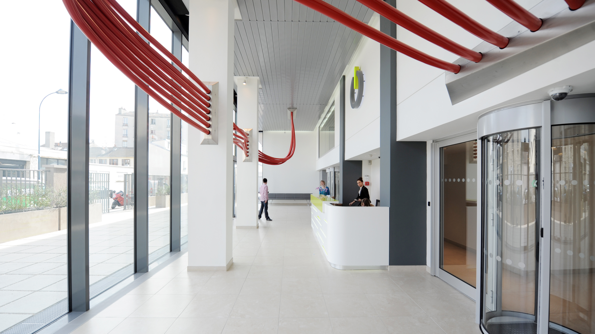Bright modern lobby with red curved tubes along the ceiling, white walls, and a reception desk with staff nearby.