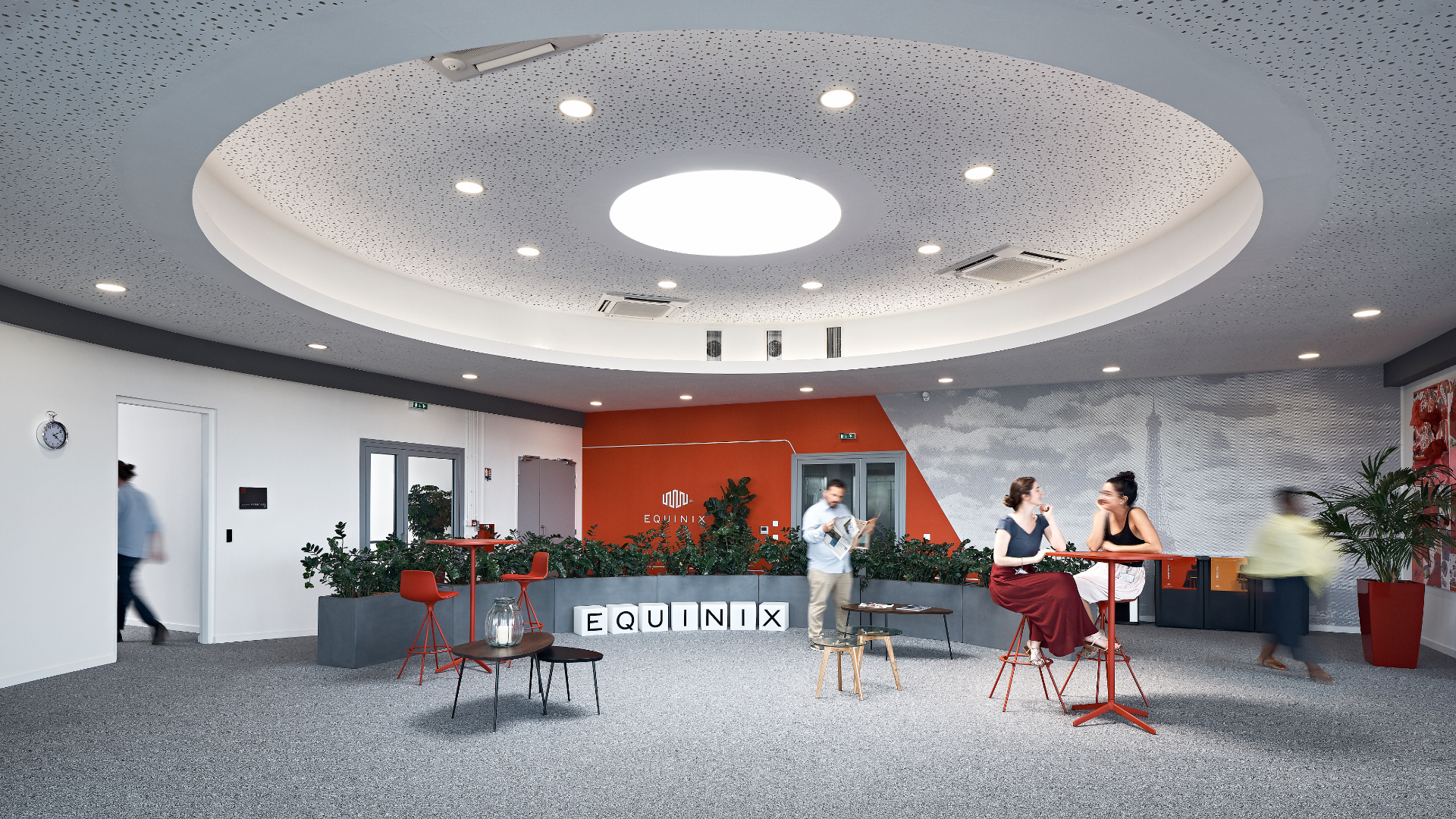 Open lobby with circular illuminated ceiling, orange wall, and people sitting at red high tables near planters and EQUNIX blocks on the floor.