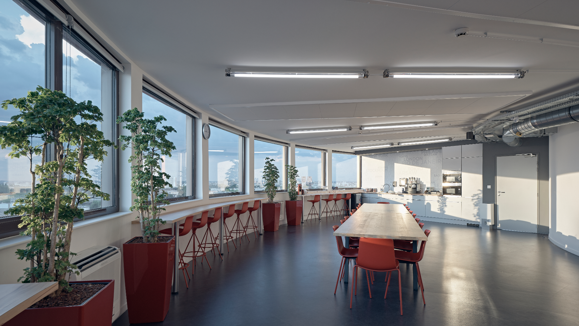 Open-plan break room with a long wooden table, red chairs, and large windows overlooking the city; tall potted plants line the walls, with a kitchenette at the far end.