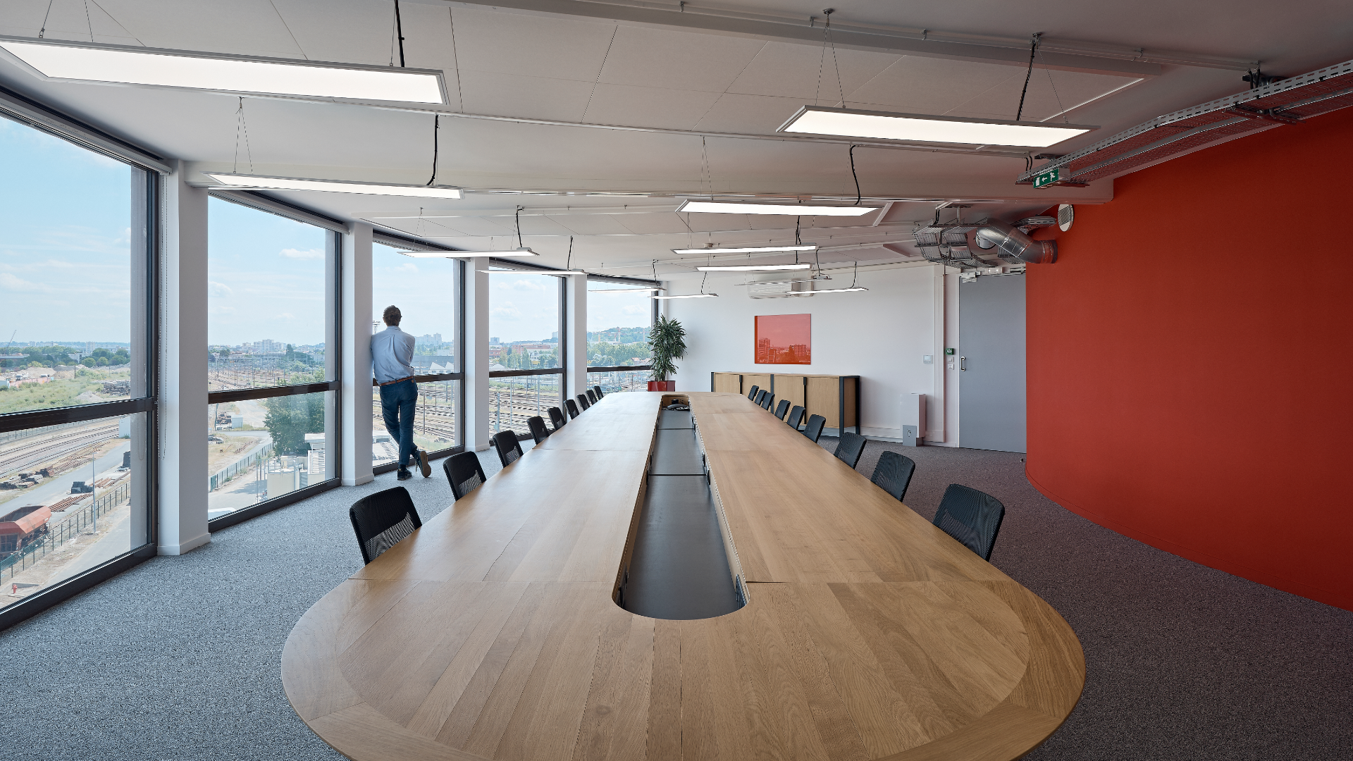 Man standing at floor-to-ceiling window in a bright modern conference room with a long curved wooden table and black chairs nearby.