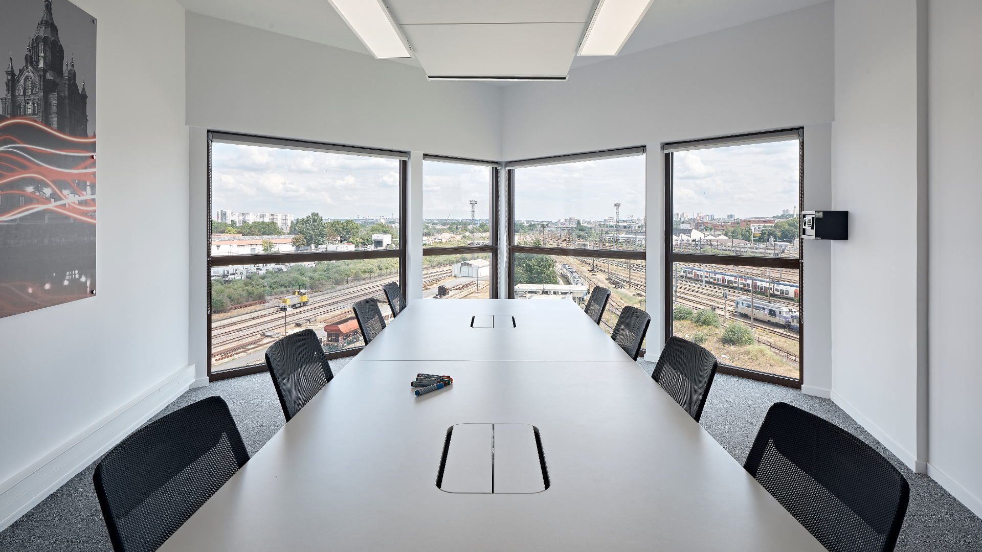 Modern conference room with a long white table and black mesh chairs, large windows overlooking railway tracks and city skyline.