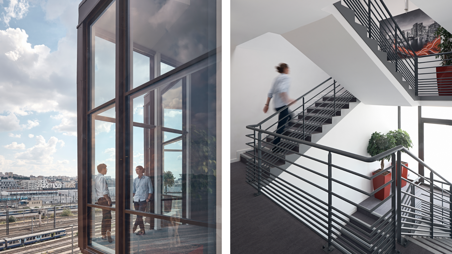 Two people in casual shirts stand and converse inside a glass-walled atrium overlooking a city skyline and rail yards.